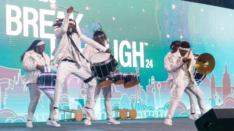Drum line performers in white uniforms energetically playing on stage at "BREAKTHROUGH 24" event with turquoise backdrop.